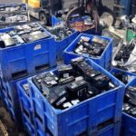 Home 12 Heavy-duty blue industrial recycling bins stacked with bulk corporate UPS batteries and scrap lead-acid power cells at a warehouse yard