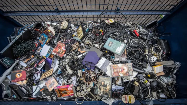 A large commercial metal bin filled with mixed electronic scrap, tangled wires, power adapters, and obsolete office devices